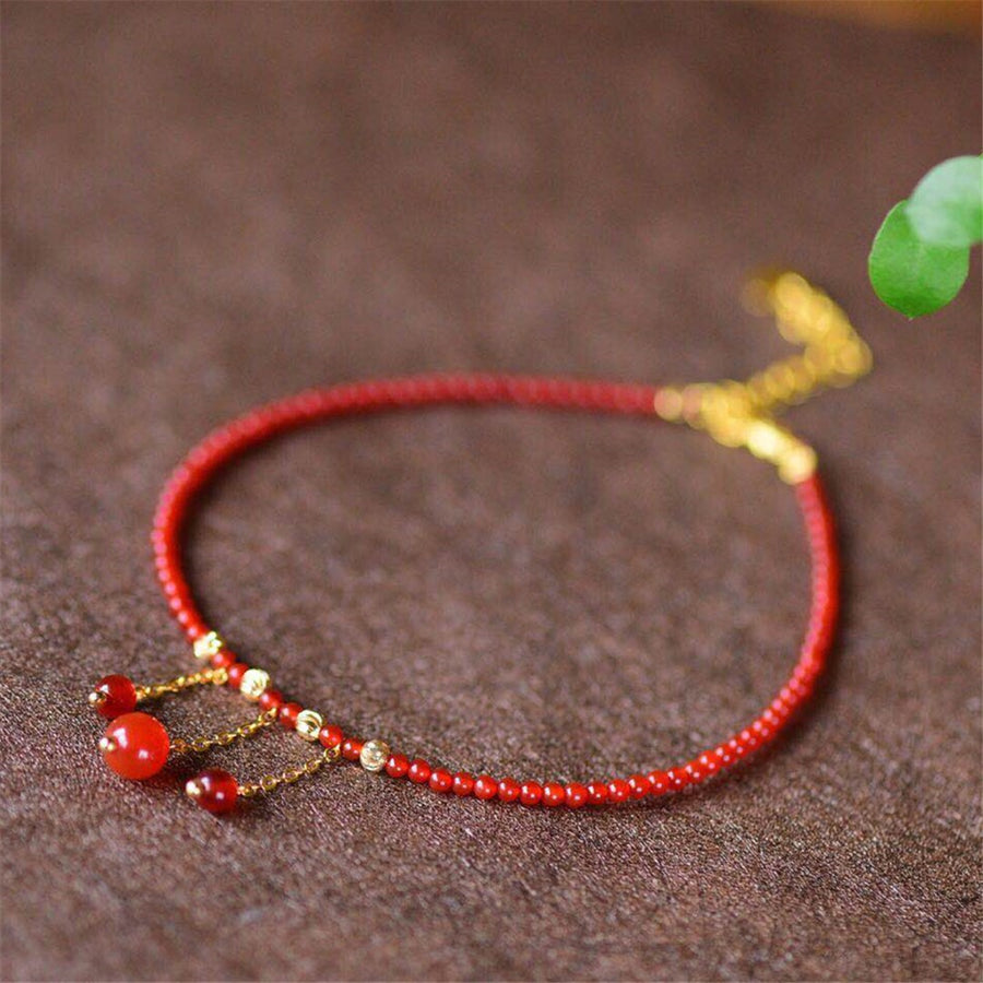 Close up of a red agate beaded anklet with gold tone tassel charms on a brown surface
