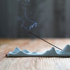 A long ceramic incense holder shaped like rolling blue-green mountains with a stick of incense burning.