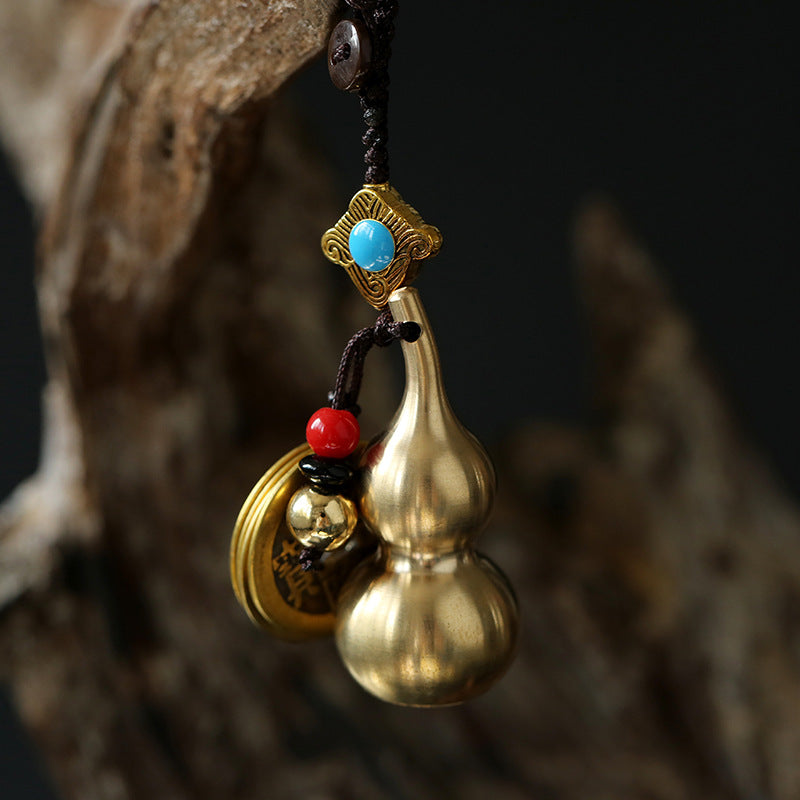 Close up of a brass gourd keychain with red beads and coins hanging from a dark cord.