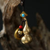 Close up of a brass gourd keychain with red beads and coins hanging from a dark cord.