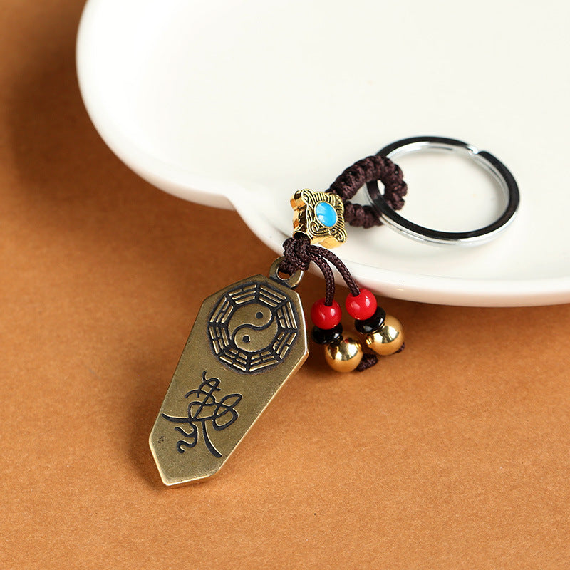 Close-up of a brass Bagua token keychain with red beads and a brown knotted cord.