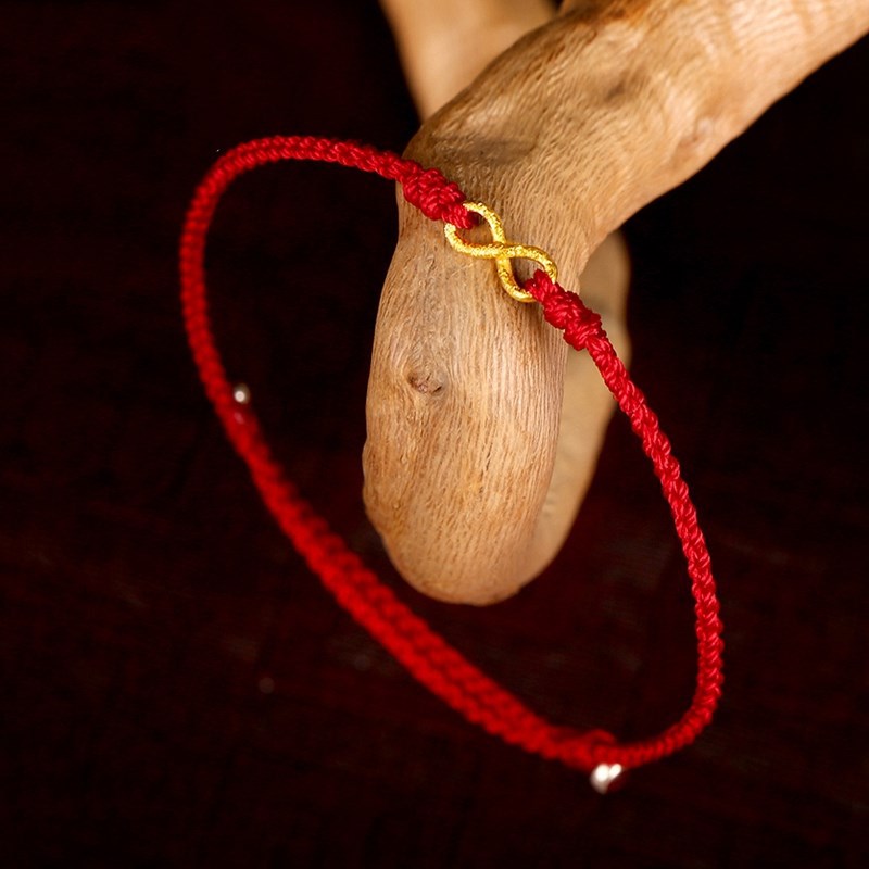 Close up of a red string bracelet with a gold infinity charm on a wooden surface