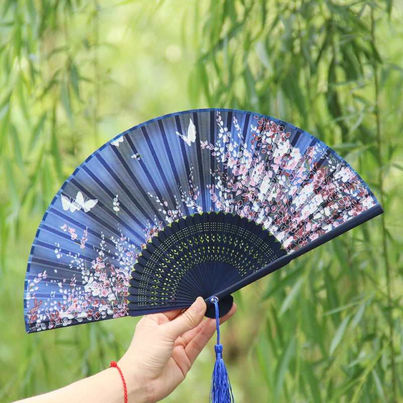 A hand holding an open blue folding fan with white butterflies and pink flowers against a green background
