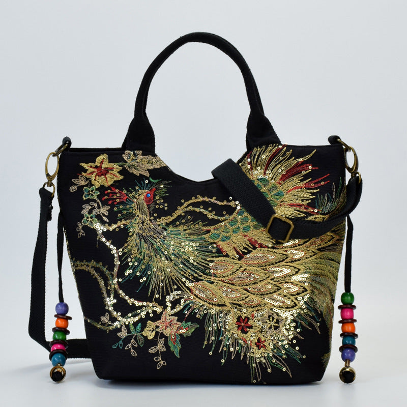 Black linen tote bag featuring gold and green embroidered peacock design with beaded strap accents