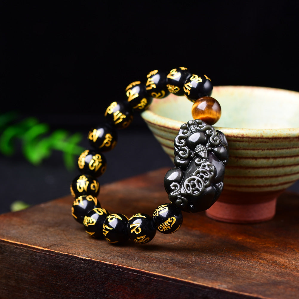 Black obsidian bracelet with carved pixiu charm and gold mantra beads on wooden surface