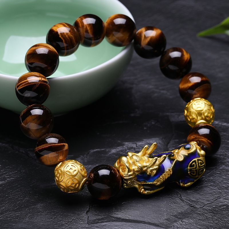 Close up of a brown Tiger Eye bead bracelet with a gold Pixiu charm resting on a dark stone surface