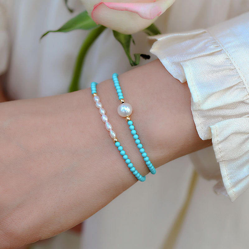 A wrist wearing two thin bracelets made of turquoise beads and white pearls.