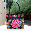 A black canvas tote bag featuring colorful double-sided embroidery of pink peonies and birds with wooden beads on the handles.