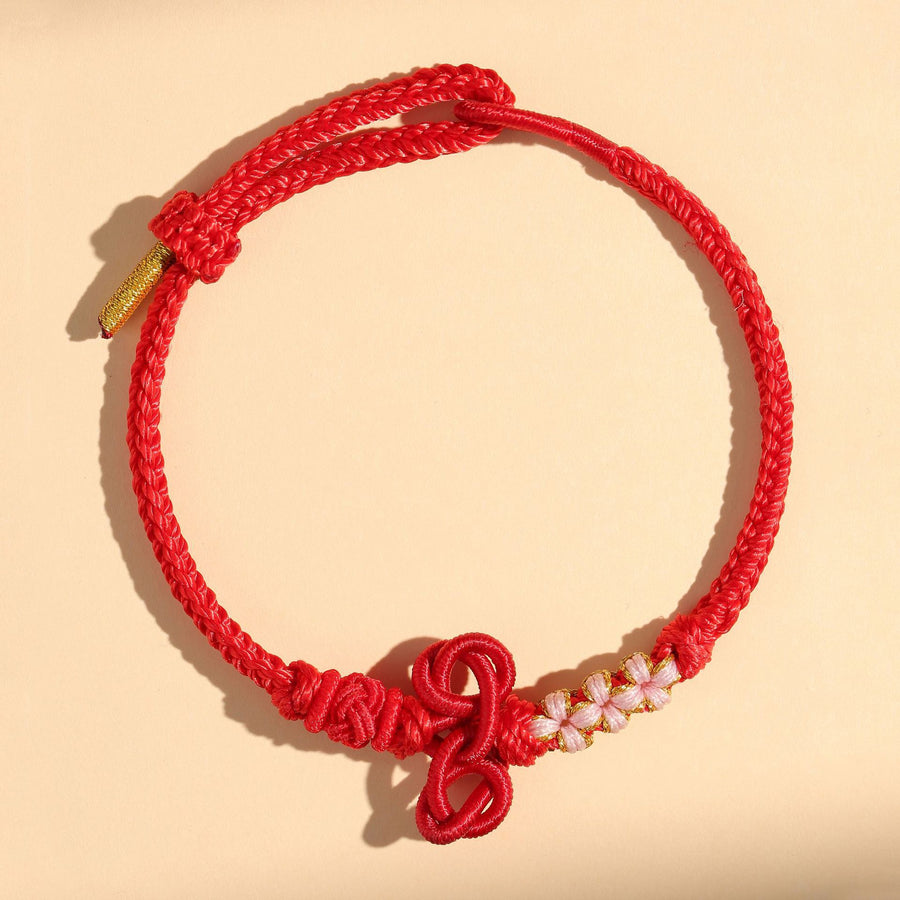 A close-up of a red braided cord bracelet with a decorative knot and white beads.