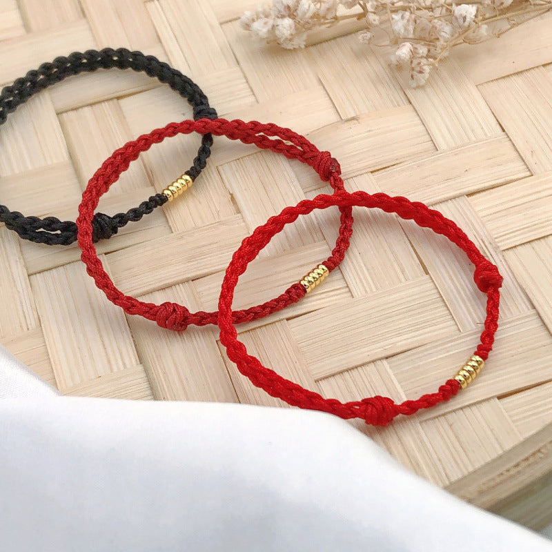 Three woven cord bracelets in red and black with a gold bead on a bamboo mat