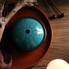 Teal mini steel tongue drum resting on a wooden tray with mallets nearby