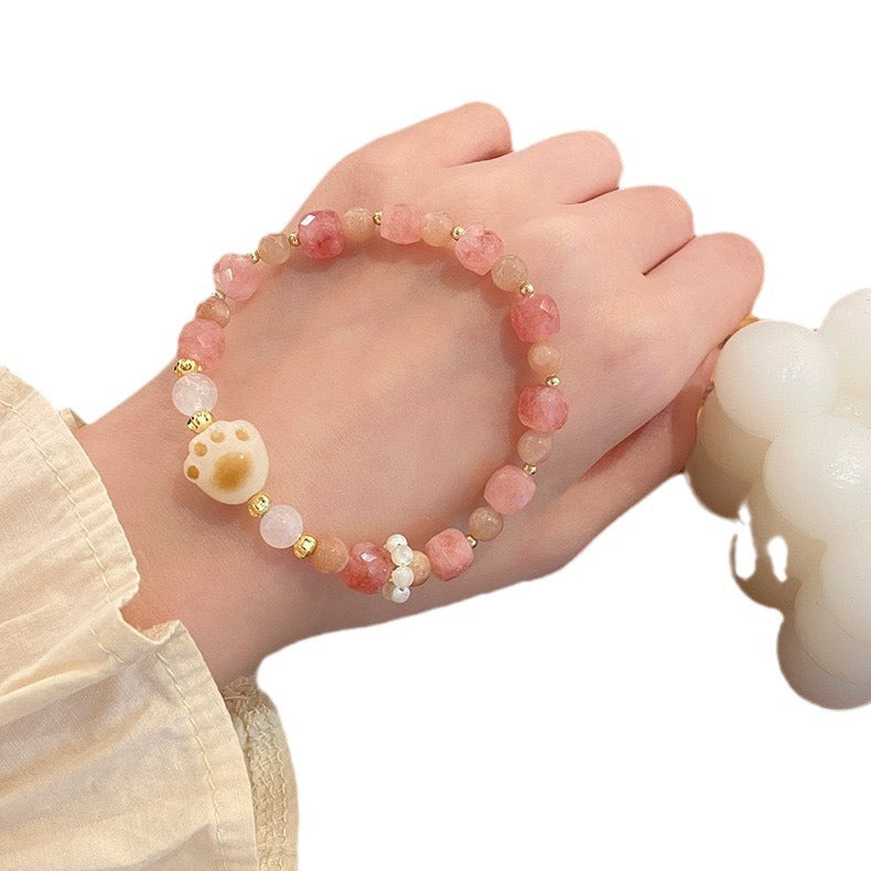 A wrist wearing a pink beaded bracelet with a white cat paw charm and gold accents.