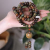 Hand holding a coiled 108 bead mala necklace with brown and green bodhi seeds and a gold pendant.