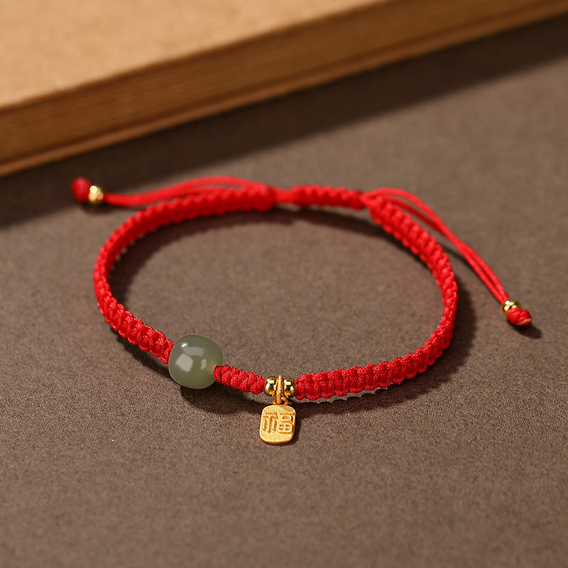 Close up of a wrist wearing a red string bracelet with a small green jade bead and gold charm