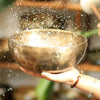 A hand holding a golden singing bowl with water droplets splashing around it