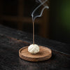 White ceramic lotus incense holder sitting on a round wooden base with smoke rising from a stick