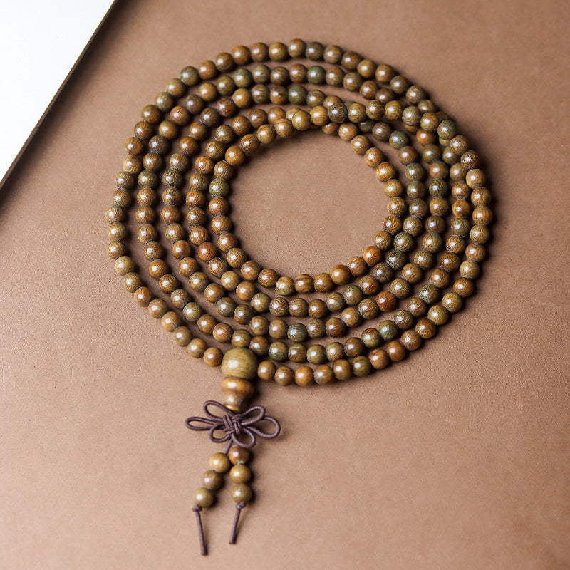 A coiled green sandalwood mala necklace with a knotted tassel resting on a beige surface