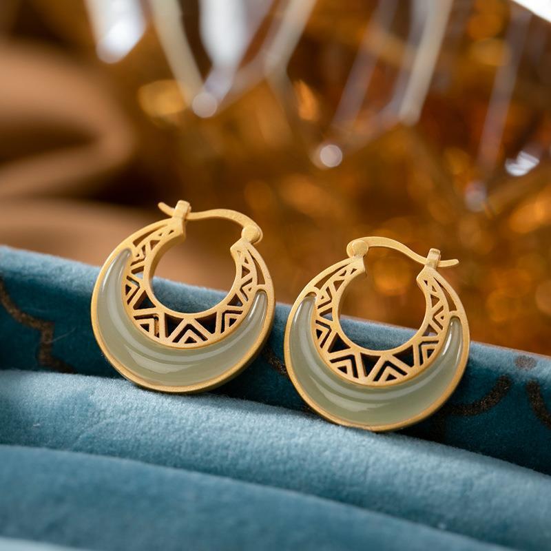 A pair of gold tone crescent moon hoop earrings with a light green faux jade curve resting on blue velvet fabric.