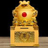 Front view of a gold-tone resin money frog statue on a box with a red gem detail
