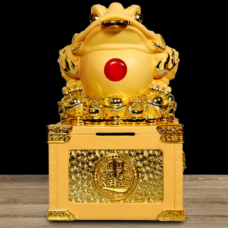 Front view of a gold-tone resin money frog statue on a box with a red gem detail