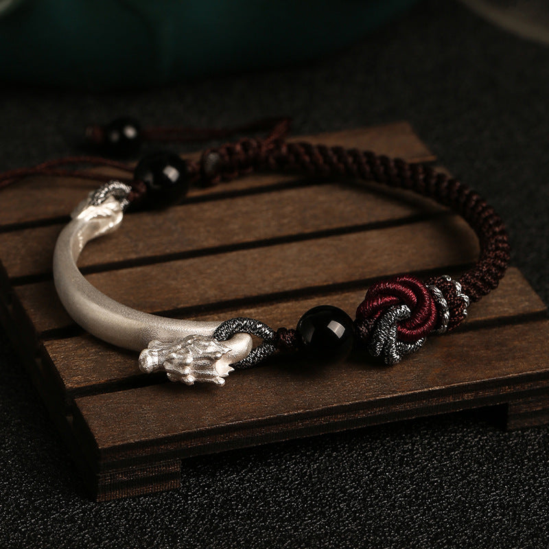 Close up of a silver dragon head bracelet on a dark brown braided cord resting on a wooden surface.