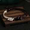 Close up of a silver dragon head bracelet on a dark brown braided cord resting on a wooden surface.