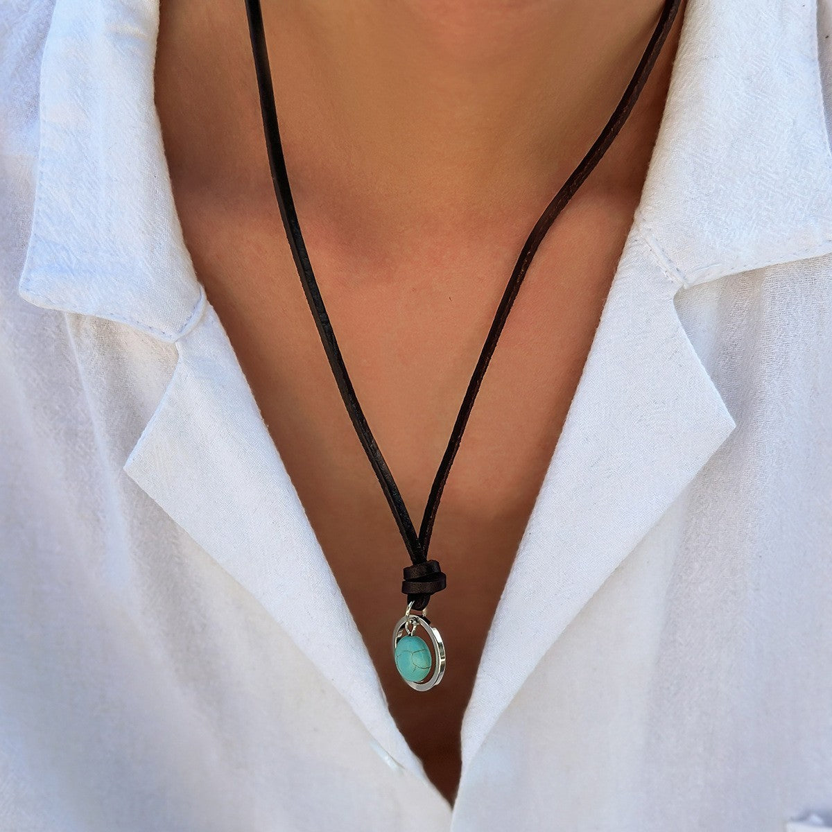 A person wearing a black cord necklace with a small round turquoise pendant against a white shirt