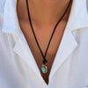 A person wearing a black cord necklace with a small round turquoise pendant against a white shirt