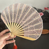 A hand holding an open folding fan with pink lace and floral embroidery