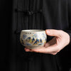 A hand holding a yellow glazed ceramic teacup painted with blue and green mountains