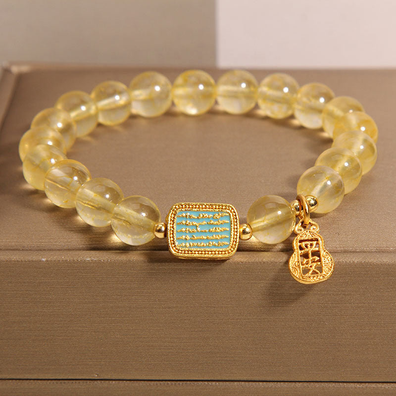 Close-up of a yellow citrine bead bracelet with a gold scripture charm on a box.