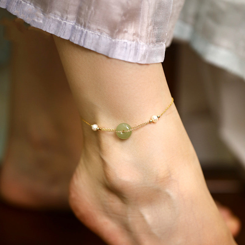 Close up of a gold tone anklet with a green jade peace buckle worn on an ankle