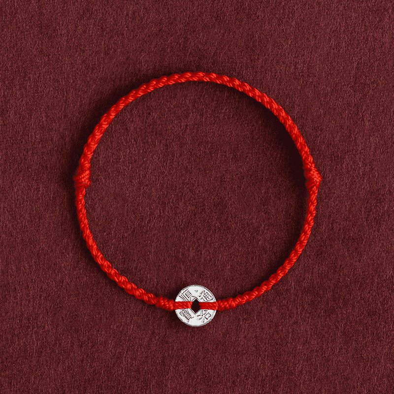 Close up of a red braided bracelet with a silver coin charm on a dark background