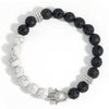 A wrist wearing a bracelet with black lava rocks, white howlite beads, and a silver Hamsa hand charm.