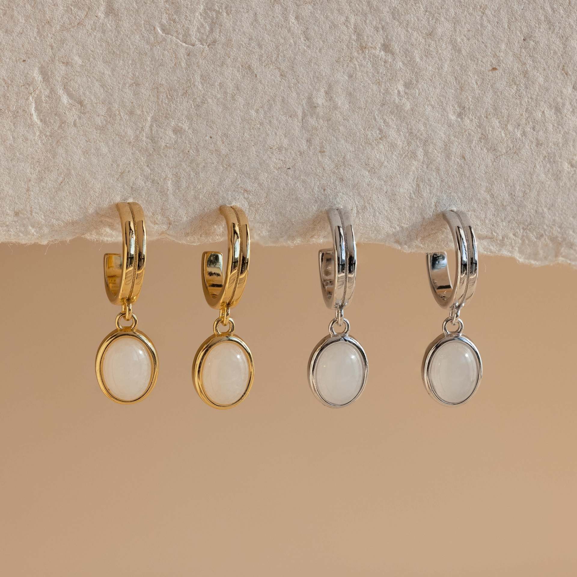Gold-tone copper hoop earrings with a white or green stone drop hanging from a textured wall