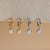 Gold-tone copper hoop earrings with a white or green stone drop hanging from a textured wall