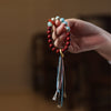 A hand holding a red cinnabar beaded bracelet with a multi-colored tassel hanging down.