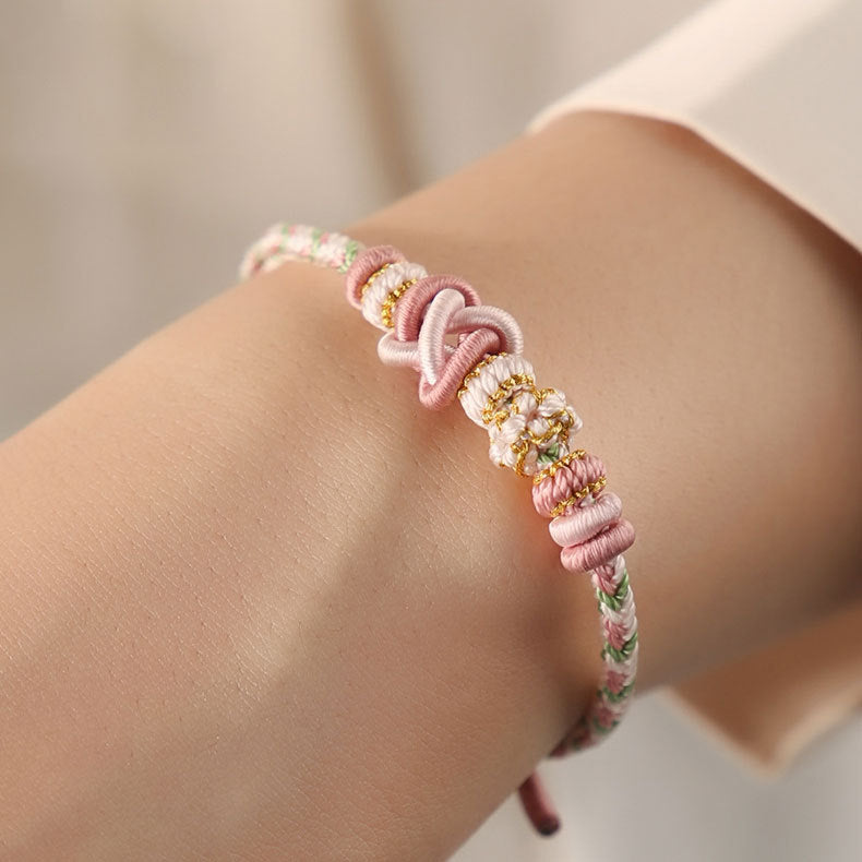 A close-up view of a pink and white woven bracelet with a gold-tone knot on a wrist.