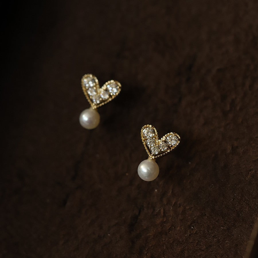 Pair of small heart-shaped stud earrings with a pearl drop on a dark background