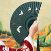 Hand holding a dark green vintage folding fan with white crane designs