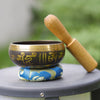 Small Tibetan singing bowl with gold mantras on a blue cushion with a wooden mallet