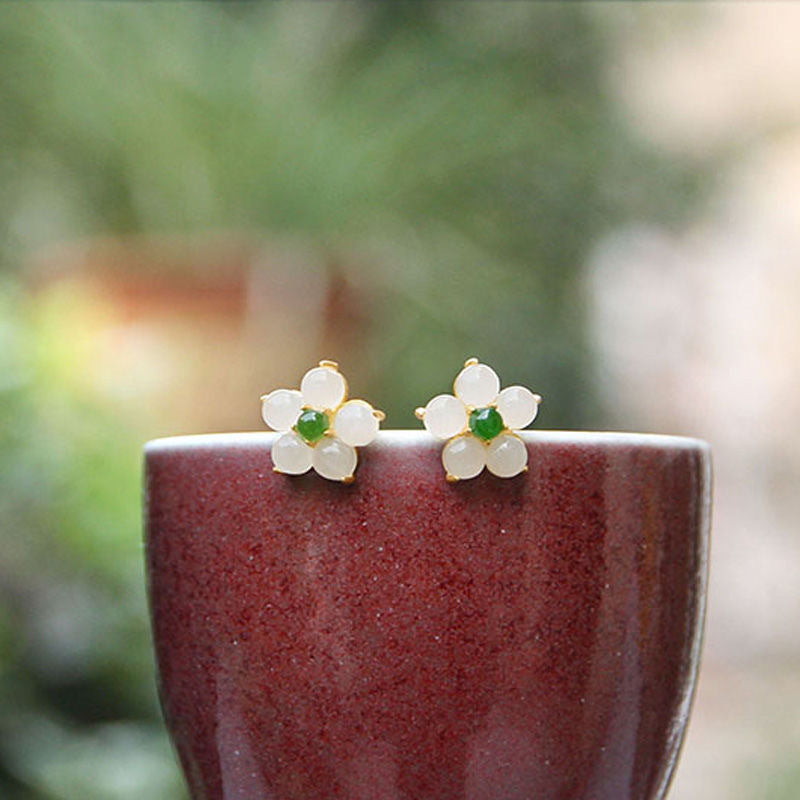 Small white jade flower stud earrings with green centers resting on a dark red ceramic cup