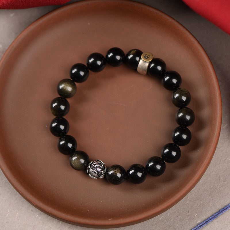 Close-up of a black and gold sheen obsidian beaded bracelet with a gold charm on a clay plate
