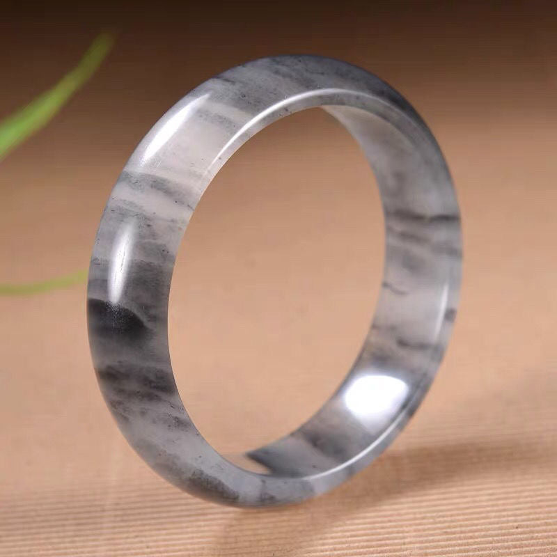 A polished gray and white golden silk jade bangle resting on a textured surface.