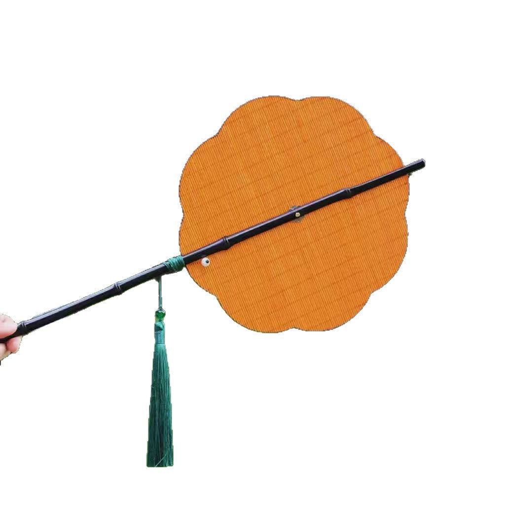 A hand holding a vintage woven bamboo fan with a long handle and green tassel against a blurred green background