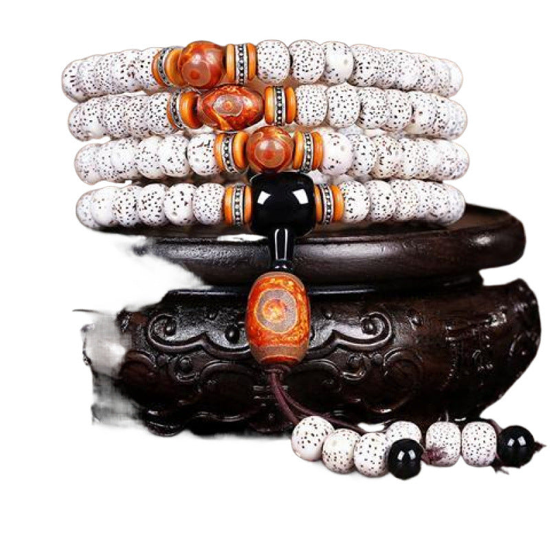 A coiled bracelet made of white speckled bodhi seeds with orange and black accent beads resting on a wooden stand.