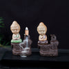 Two ceramic incense burners with Buddha figures, one green and one brown, showing backflow smoke.