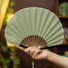 A hand holding an open light green shell shaped folding fan with a bamboo frame