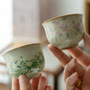 Two hands holding small ceramic tea cups with pink flower and white daisy designs