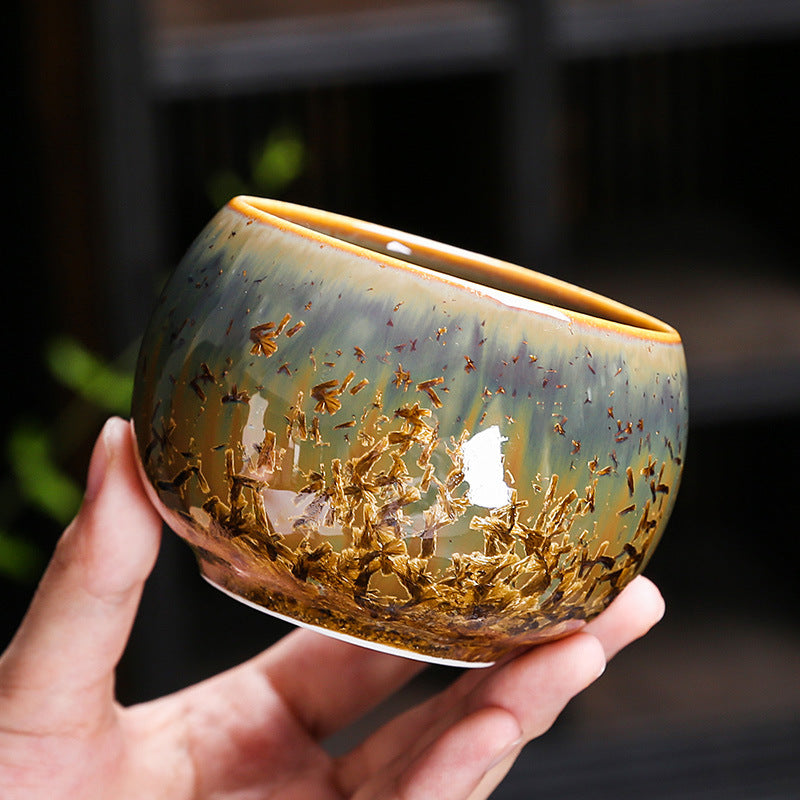 Hand holding a round ceramic tea cup with gold-tone and green crystalline glaze patterns